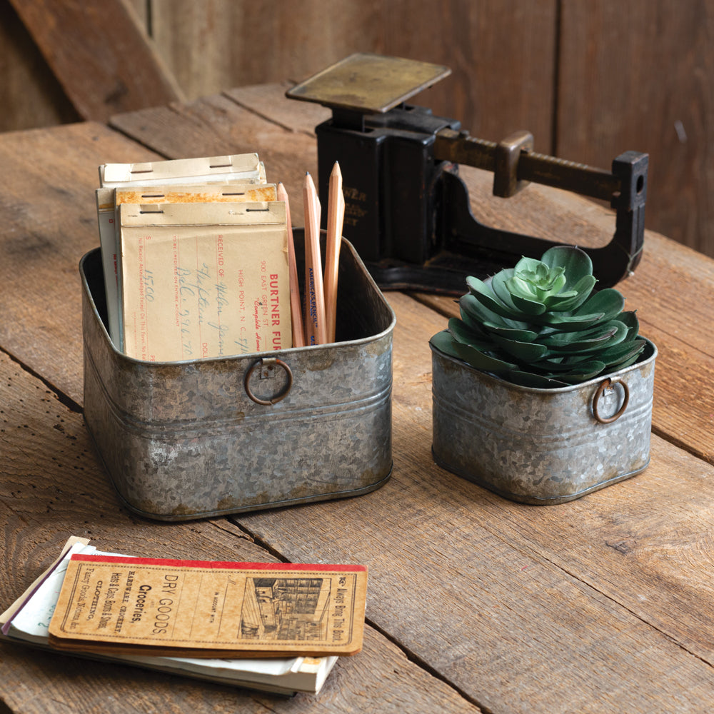 Two galvanized square bins with black metal handles, one larger and one smaller, showcasing rustic elegance on a wooden tabletop.