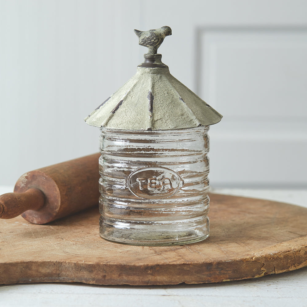 Clear glass canister with a rustic metal lid featuring a bird, adorned with a tea emblem, perfect for tea lovers and collectors.